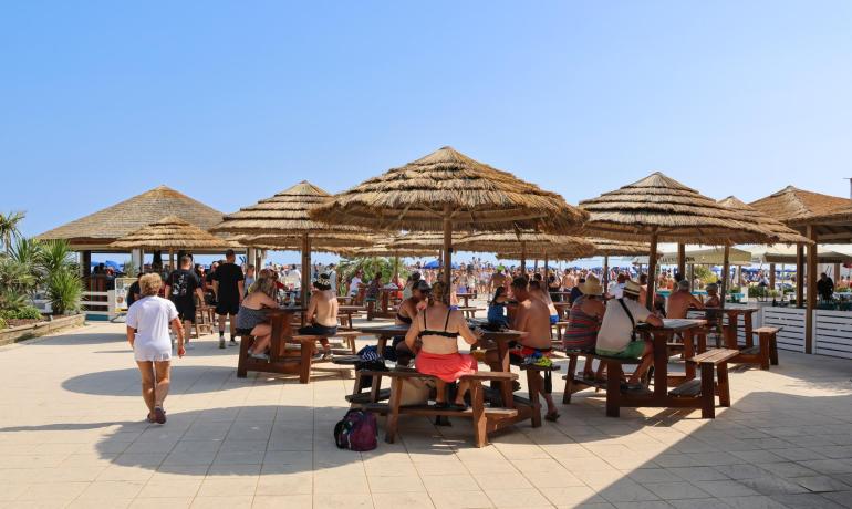 Crowded beach with straw umbrellas and wooden tables.