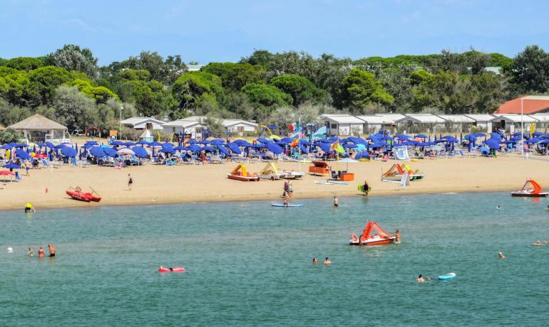 Crowded beach with blue umbrellas and swimmers in the sea.