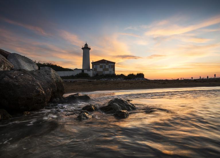 Sunset over lighthouse with sea and rocks in foreground.